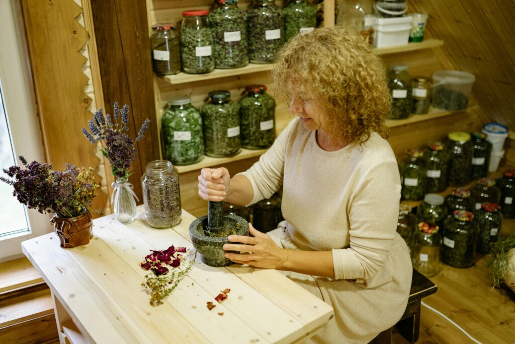 A woman uses a mortar and pestle to prepare herbal remedies in a rustic kitchen setting.